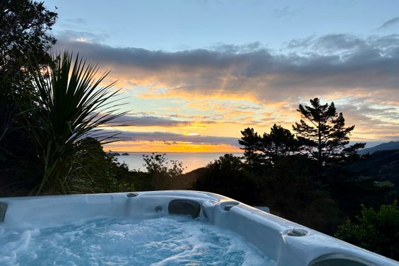 A bubbling outdoor hot tub at Ocean View Retreat looks out over a coastal sunset. Golden sunlight streams through clouds over the sea, framed by native New Zealand bush, a cabbage tree, and rolling green hills.